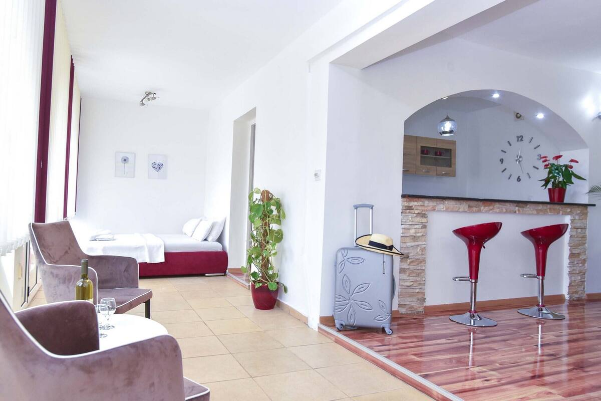 A modern hotel room with a bed, a dining area with a table and chairs, a kitchen area with a clock and red bar stools, and a potted plant.