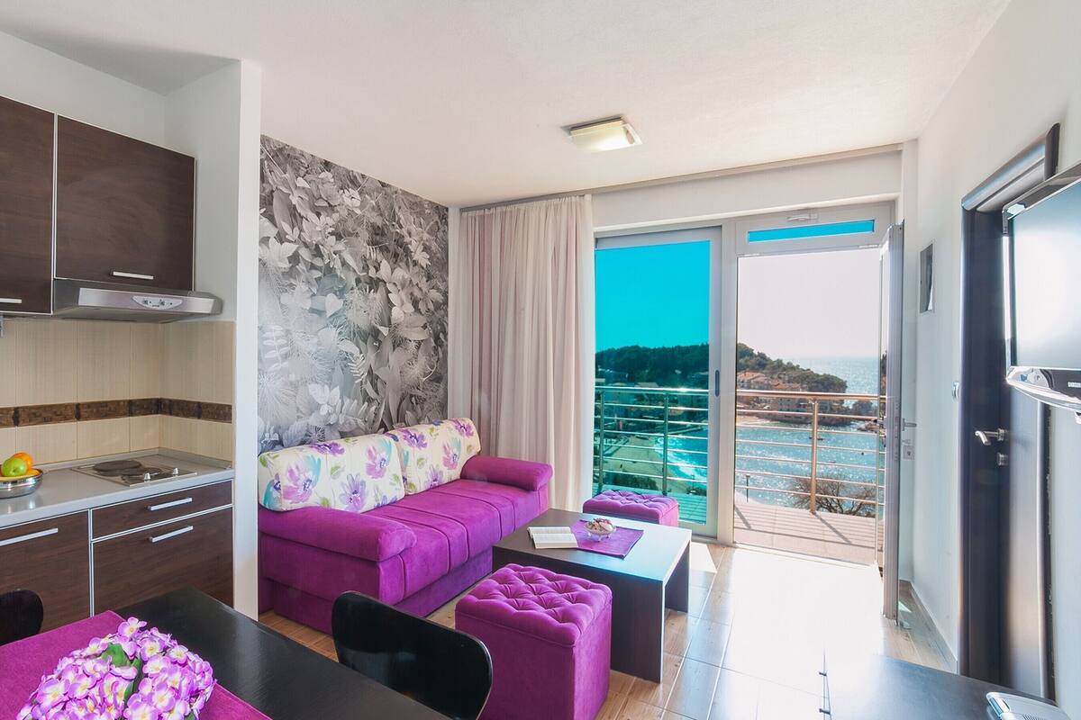 A modern kitchen with a dining area, a purple sofa, and a balcony with a view of the sea.