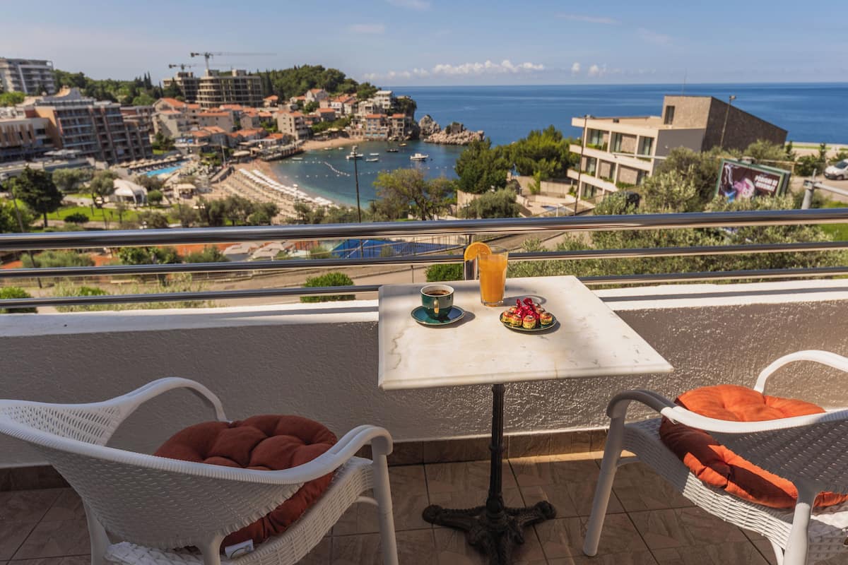 A balcony with wicker chairs, a small table with a drink and a plate of food, and a view of a beach and buildings.