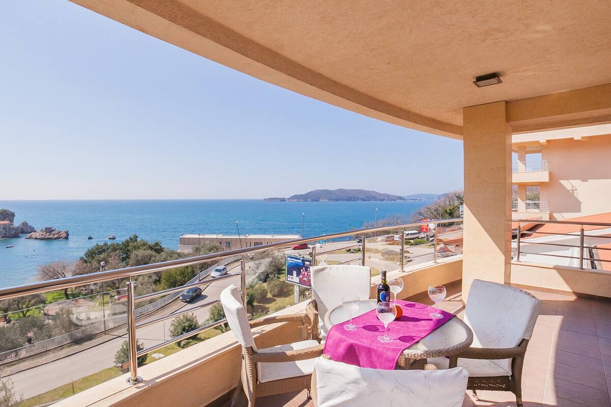 A balcony with wicker furniture, a table with a pink tablecloth, and a view of the sea and buildings.