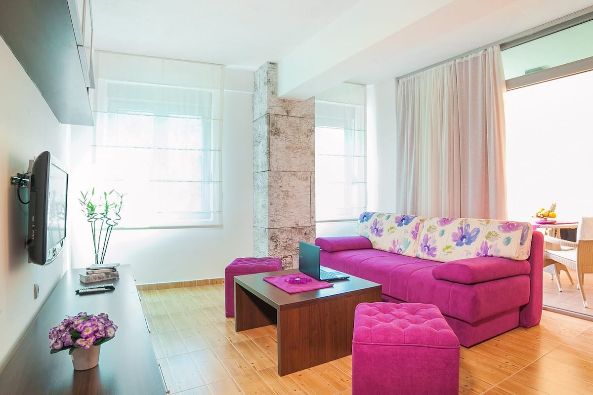 A modern living room with a pink sofa, a coffee table, and a laptop. There is a TV mounted on the wall, a small table with a vase of flowers, and a window with curtains.