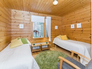 A cabin with two single beds, a small table, a lamp, and a view of a cabin through a window.