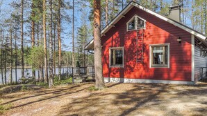 A red cabin with a wooden deck, surrounded by tall trees and a body of water. - Sammonranta by Interhome (Lieksa)