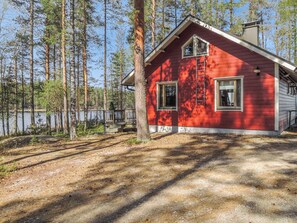 A red cabin with a wooden deck, surrounded by tall trees and a body of water.