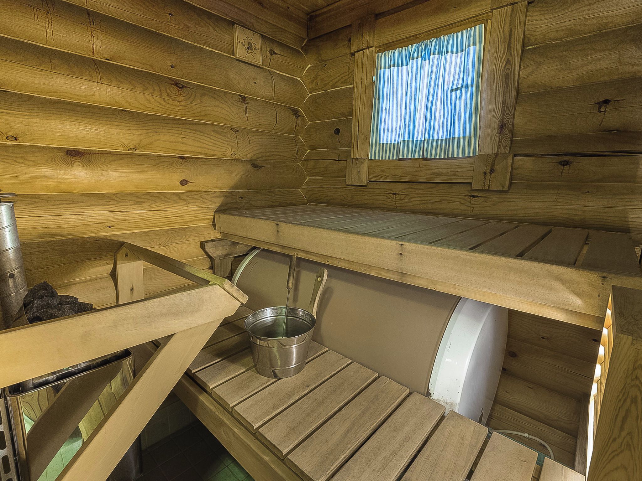 L’intérieur d’une petite cabane en bois est aménagé avec un lit superposé, une petite fenêtre avec des rideaux et un seau en métal posé sur le sol.