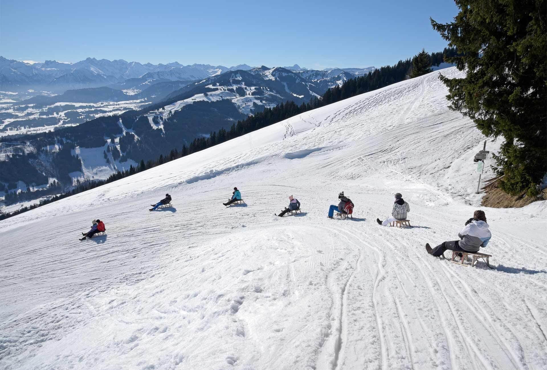 Ein verschneiter Skipisten mit Personen, die auf Schlitten sitzen.