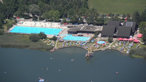 Vista aérea de un resort con dos piscinas grandes, un puente que las conecta y un edificio con techo oscuro.