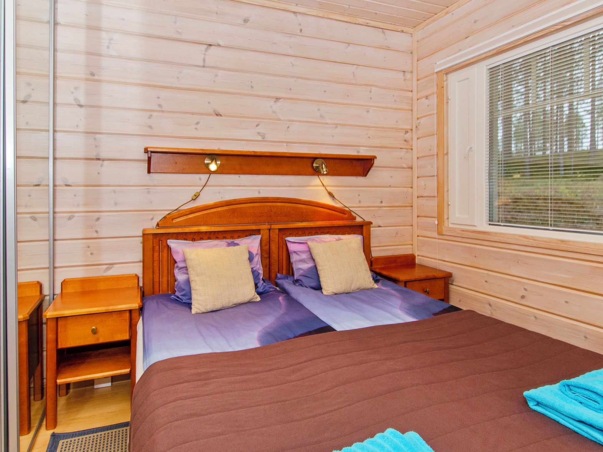 Ein Zimmer in einer Holzhütte mit einem Bett, zwei Nachttischen, einem Fenster mit Jalousien und einem Holzkopfende mit Beleuchtung.