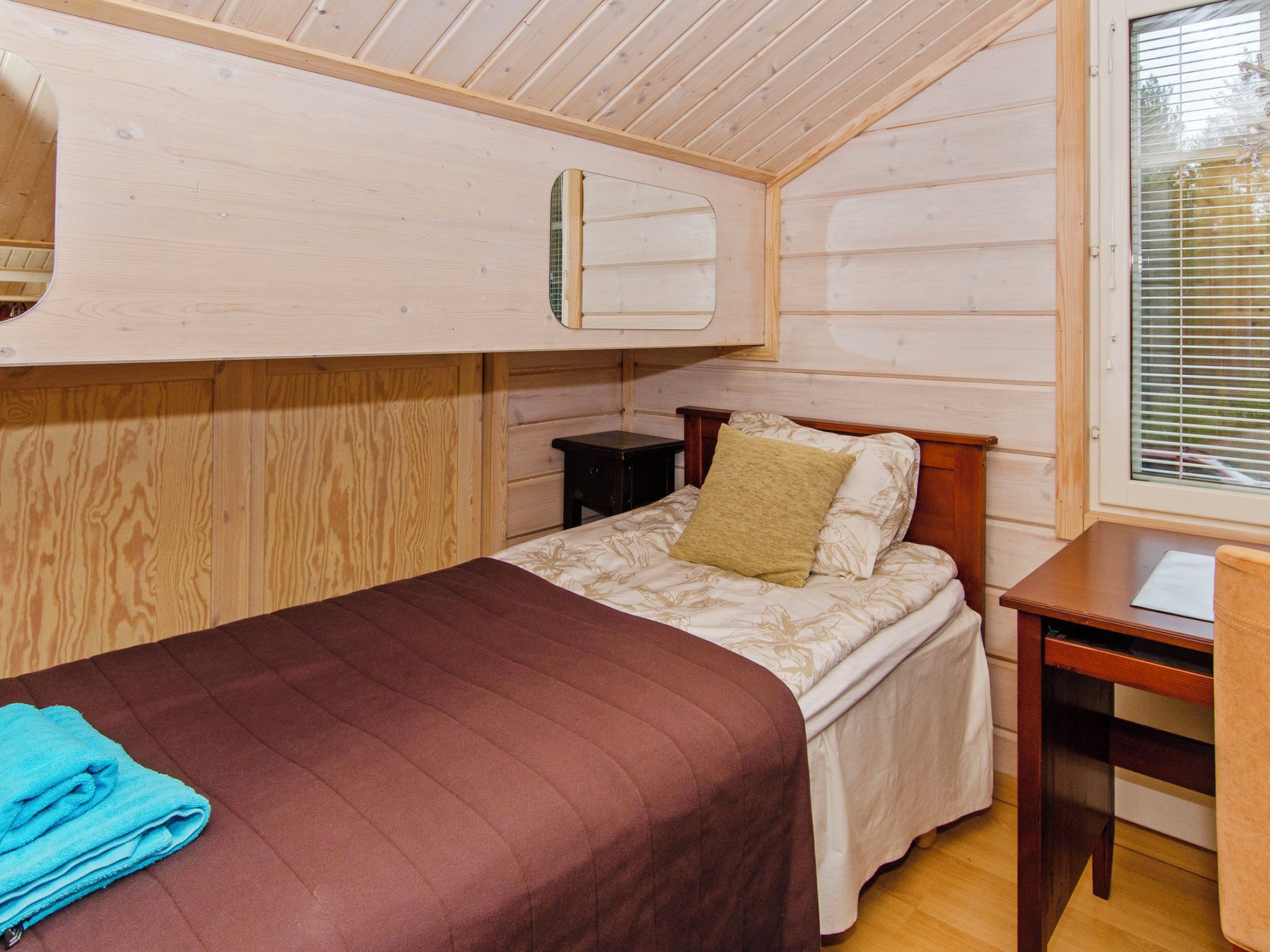 A single bed with a brown bedspread, a bedside table, a small nightstand, and a window with blinds.
