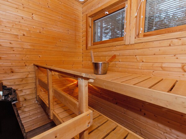 A wooden cabin interior with a wooden bench, a metal bucket, and a window with blinds.