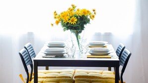 Mesa de jantar posta para seis pessoas, com um vaso de flores amarelas, copos de vidro transparente e cadeiras com almofadas amarelas.