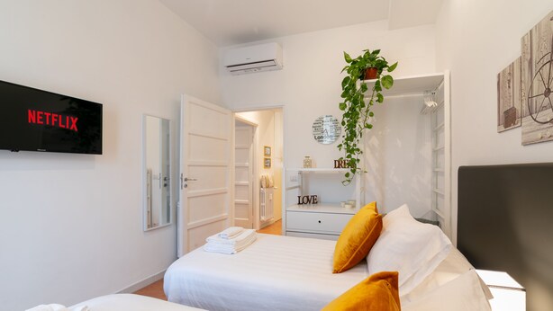 Quarto de hotel com duas camas, uma televisão exibindo 'NETFLIX', uma planta e um letreiro com a palavra 'LOVE' sobre um penteadeira.