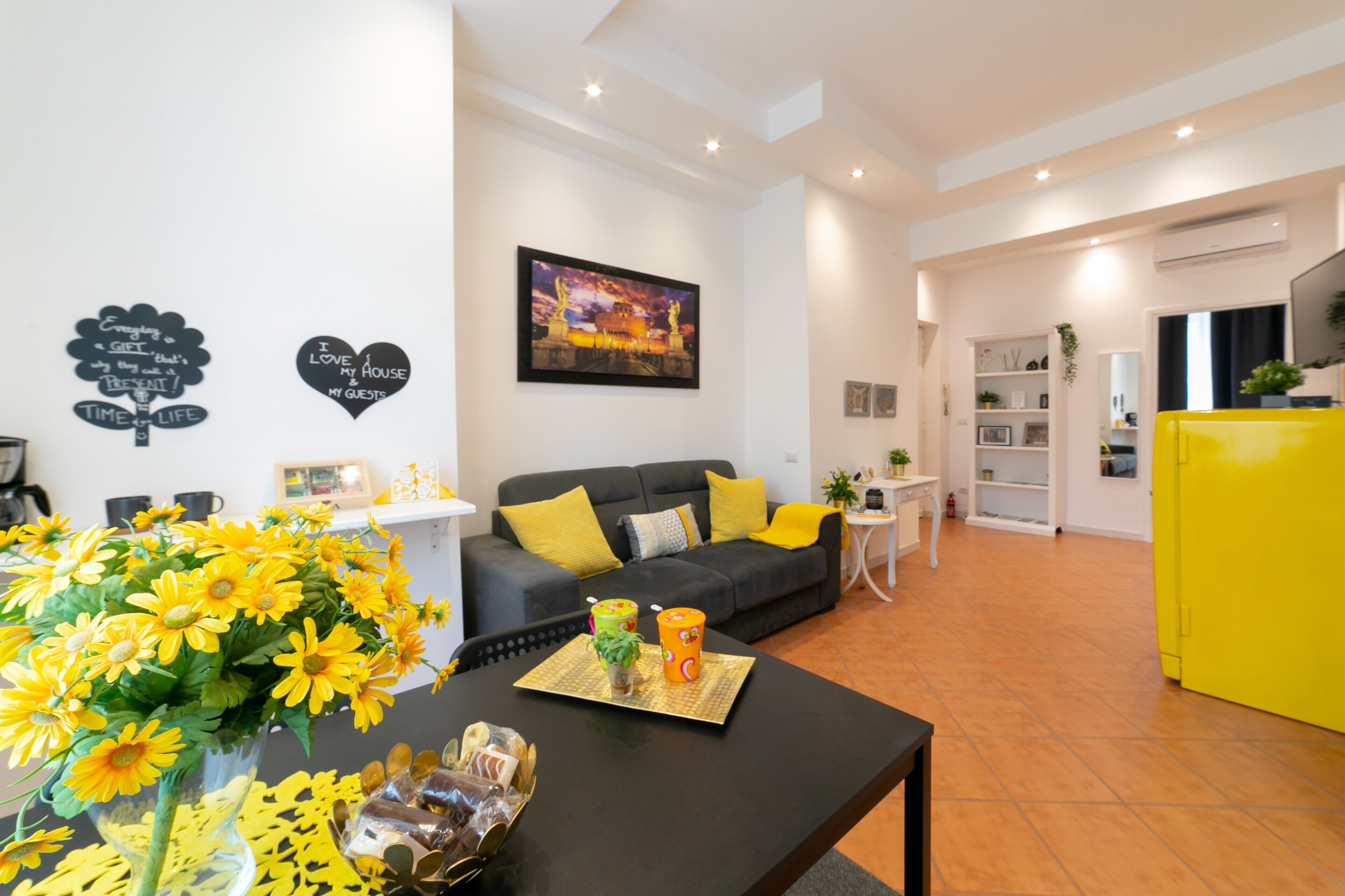 Una sala de estar moderna con un sofá, una mesa de centro y un mueble amarillo. Hay objetos decorativos en las paredes y un cuadro sobre el sofá.