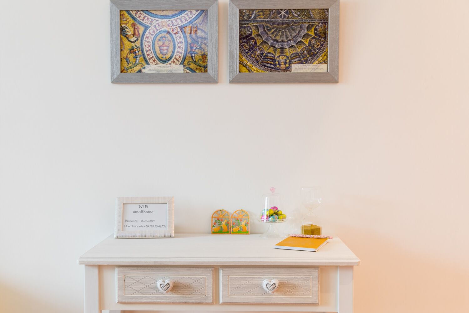 Une commode blanche ornée de deux œuvres d’art encadrées, d’une pancarte indiquant le mot de passe du Wi-Fi, ainsi que de divers objets décoratifs, notamment un bocal en verre rempli de bonbons colorés et une petite figurine orange.