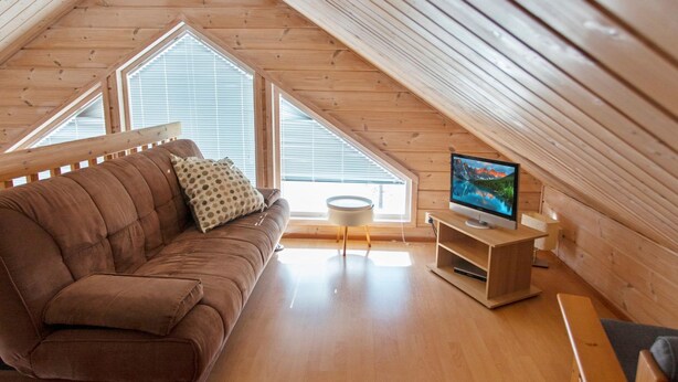 A cozy attic room with a brown sofa, a television on a wooden stand, and large windows allowing natural light.