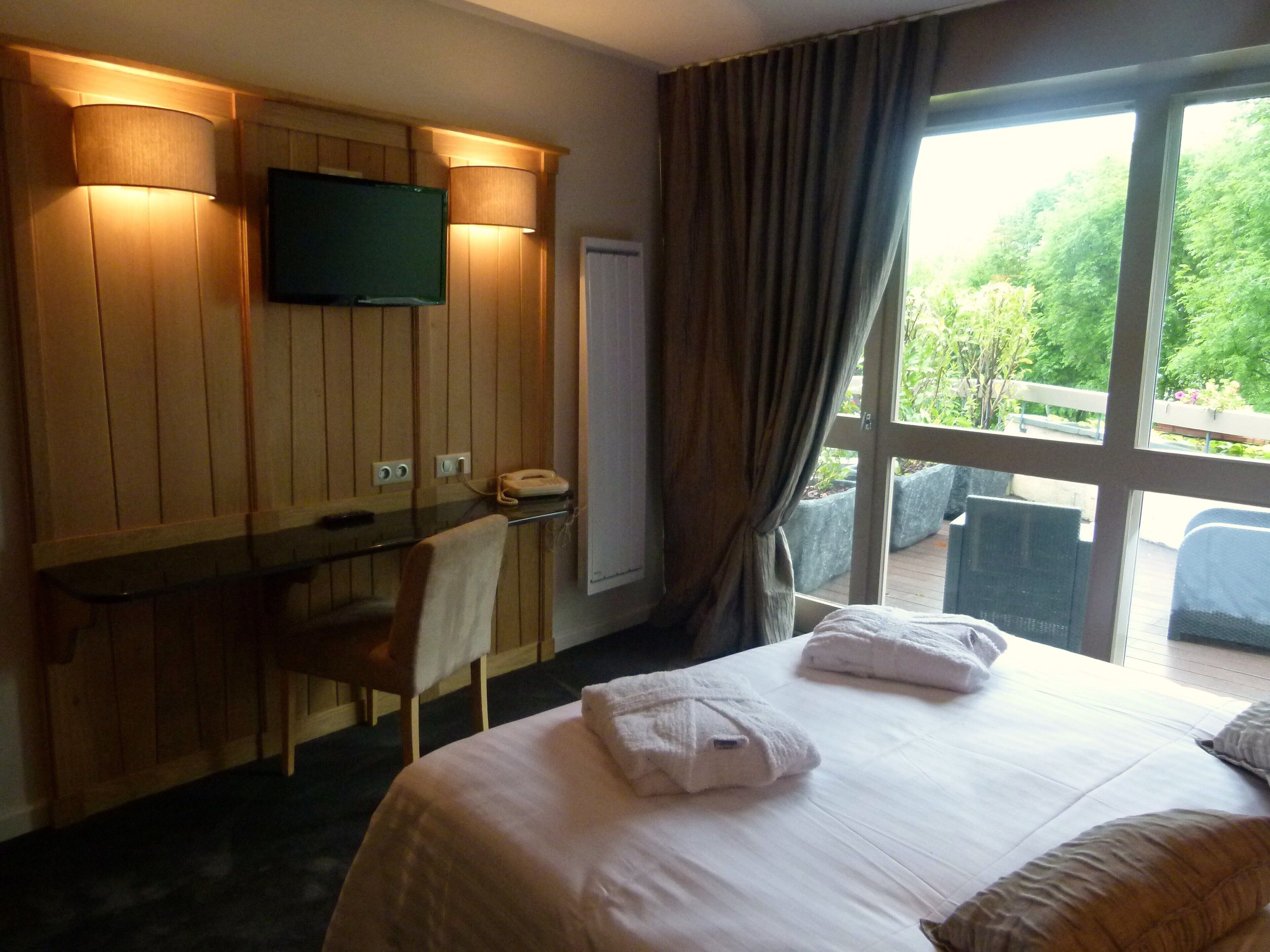 A hotel room with a bed, a desk, a chair, a TV mounted on the wall, and a window with curtains.
