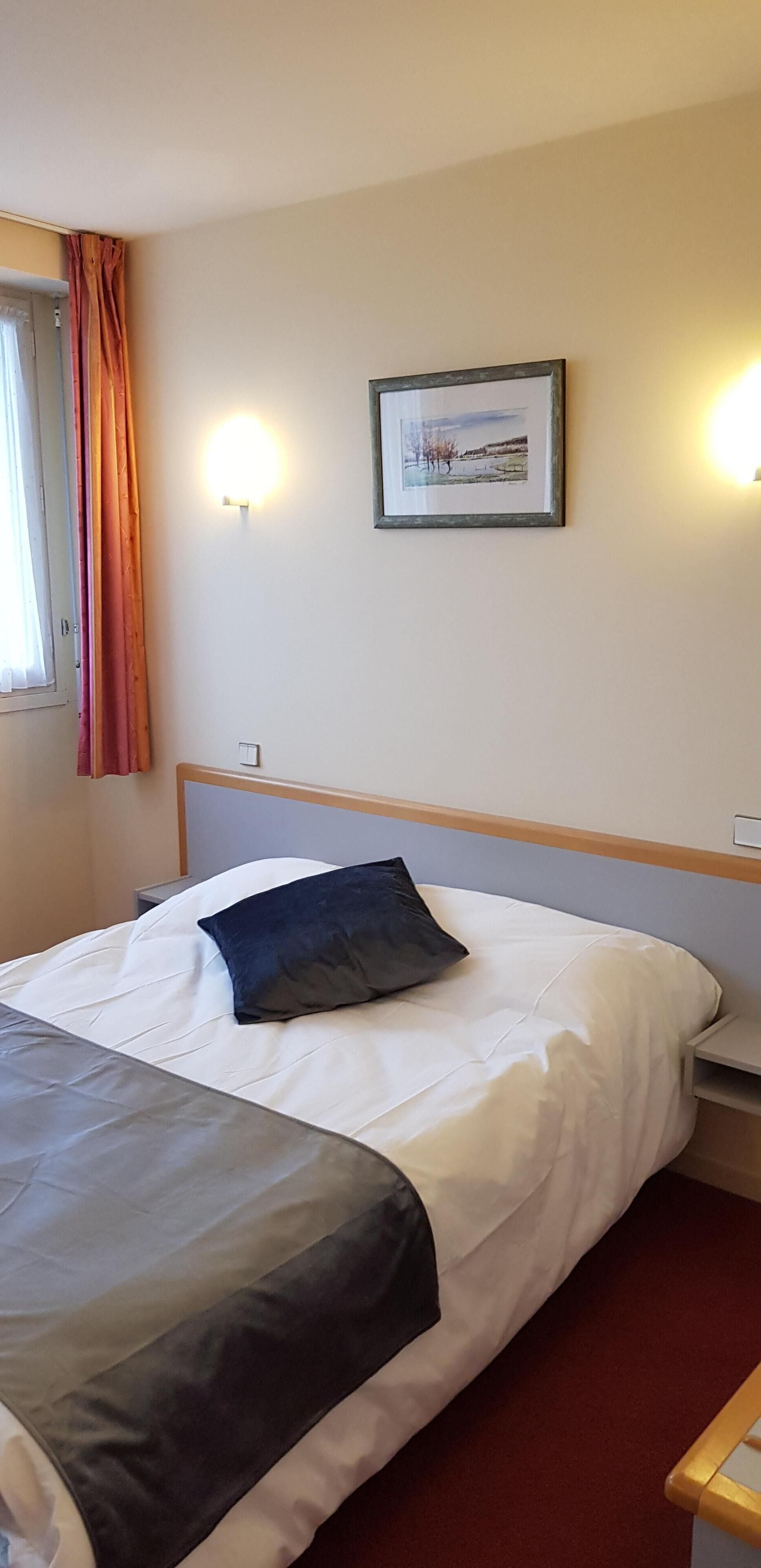 A hotel room with a bed, a nightstand, a wall-mounted lamp, a window with curtains, and a framed picture on the wall.