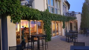 Un espace extérieur aménagé avec des tables et des chaises, un bâtiment doté d’une façade en verre et un arbre en arrière-plan.