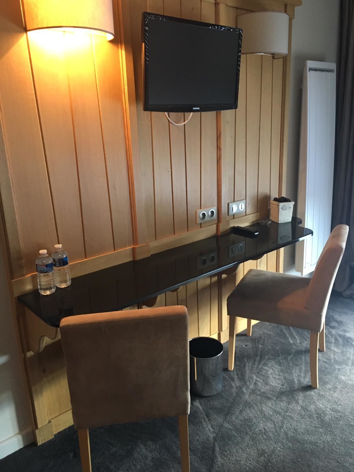 Habitación de hotel con pared revestida de madera, un televisor de pantalla plana montado sobre un escritorio de cristal, dos sillas, un cubo de basura y una botella de agua sobre el escritorio.