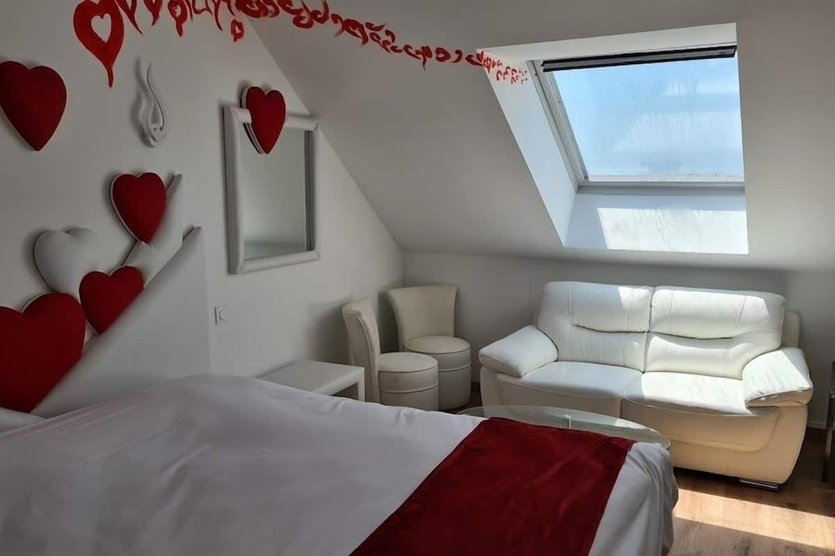 Ein modernes Schlafzimmer mit schrägem Dach, einem großen Bett mit rotem Akzent, einem weißen Sofa und einem Dachfenster.