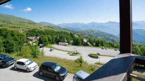 Un aparcamiento con varios coches, rodeado de vegetación y edificios, con un paisaje montañoso al fondo.