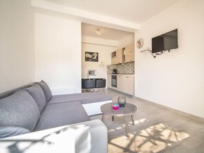 Un salon moderne avec un canapé gris, une petite table ronde sur laquelle est posé un vase décoratif, et un coin cuisine à l’arrière-plan.