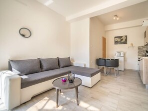 Un salon moderne avec un canapé d’angle gris, une petite table ronde et un coin cuisine équipé d’un micro-ondes et de chaises.