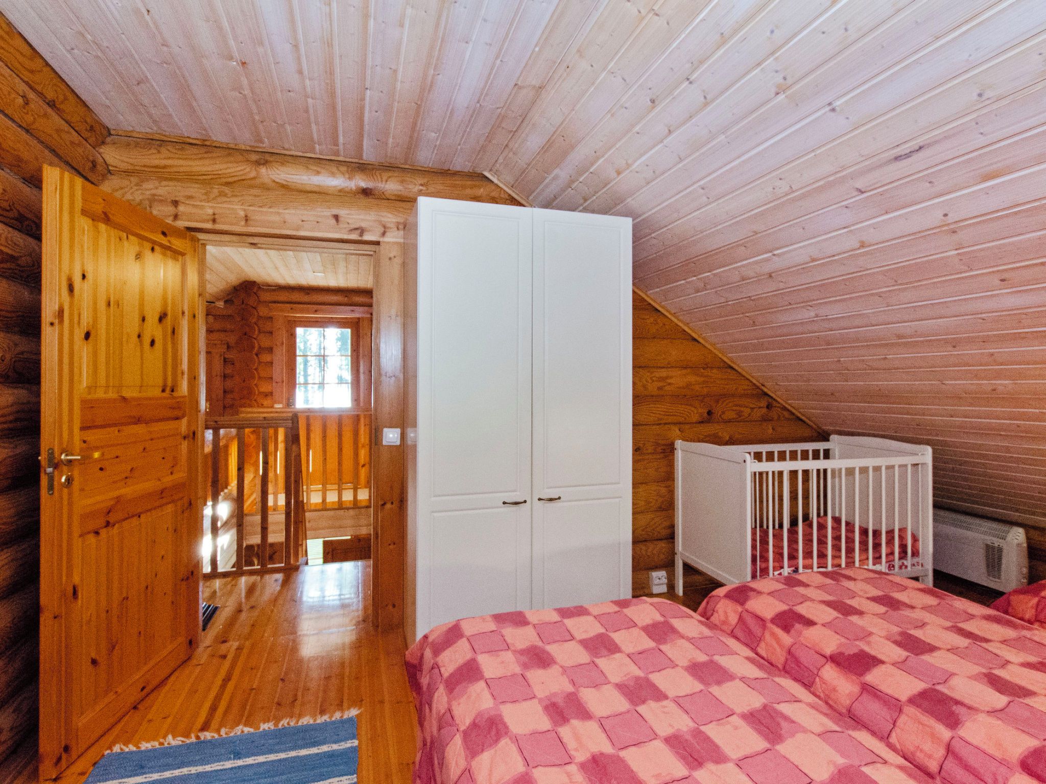 A bedroom with a wooden ceiling, a white wardrobe, a crib, and a checkered bedspread.