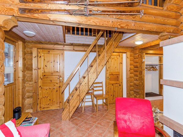 Interior de una cabaña de troncos con una escalera de madera, una silla roja y una mesa de madera con revistas.