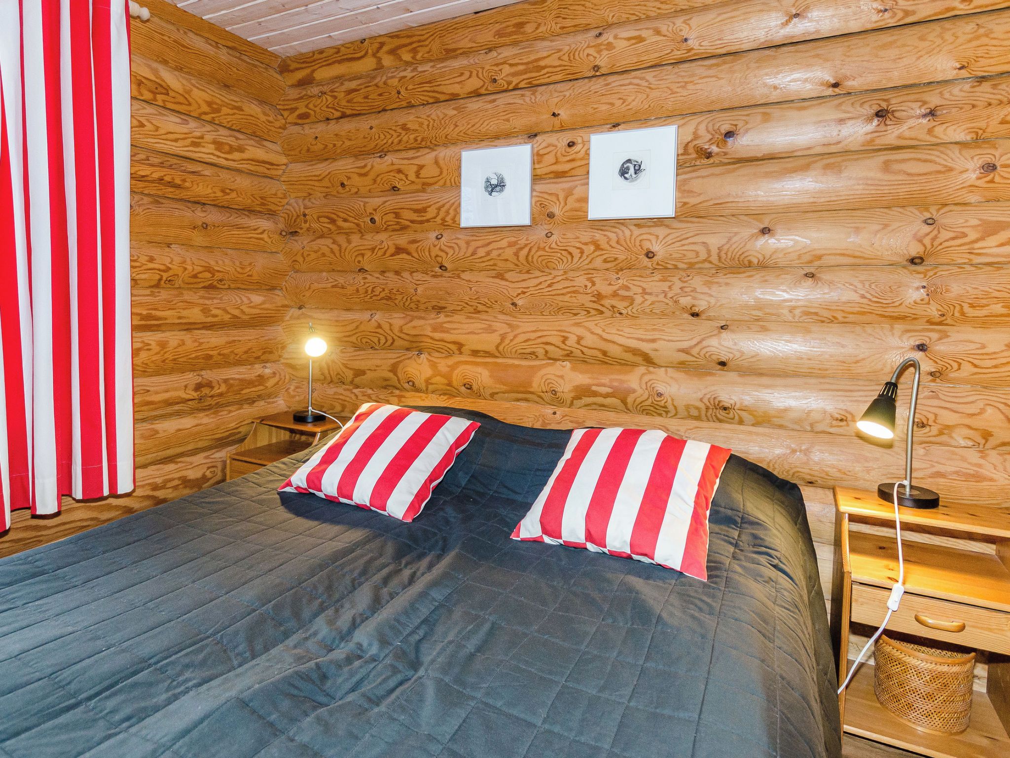 A wooden cabin room with a bed, two bedside lamps, and a red and white striped curtain.