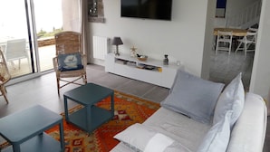 A living room with a sofa, a TV, a dining area with a table and chairs, and a sliding glass door leading to an outdoor area. - Character house with 180° panoramic sea view - Côte Sauvage de Portsall (Landunvez)
