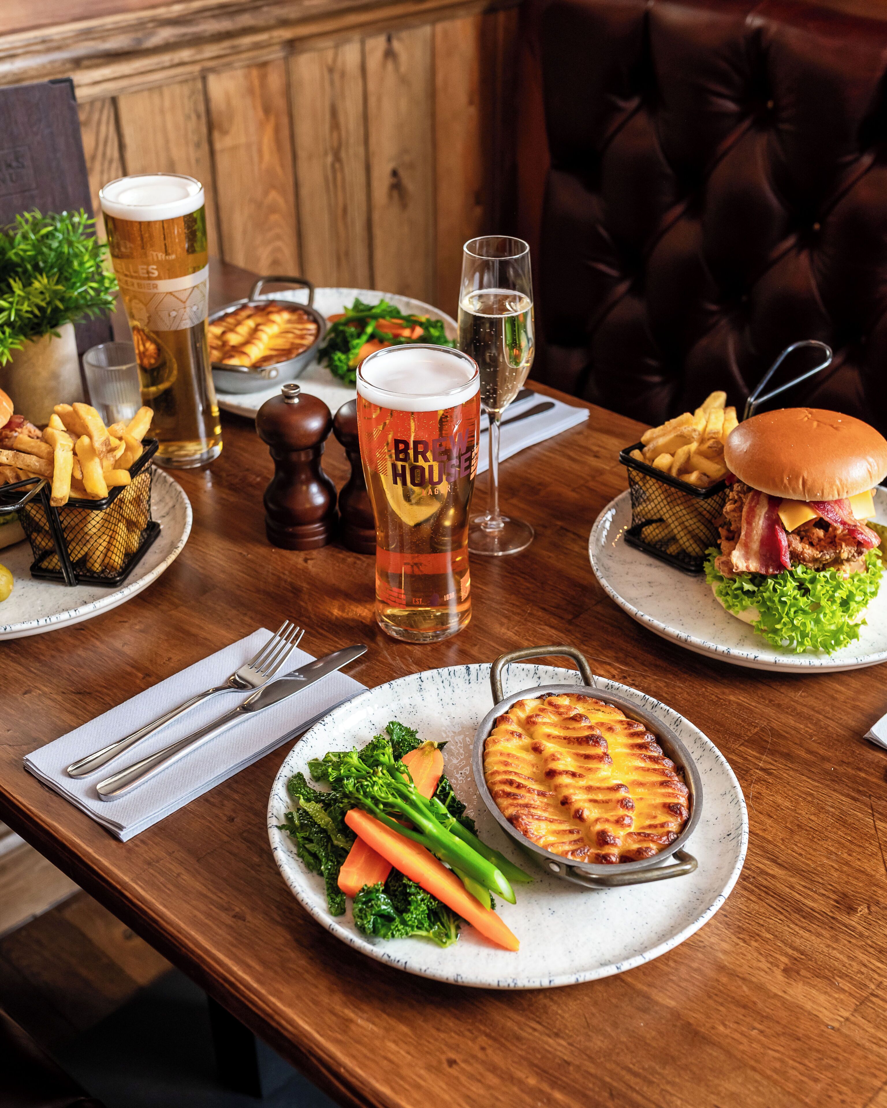 Una mesa de comedor con diversos platos, entre ellos una hamburguesa, papas fritas y una tarta, además de vasos de cerveza y una ensalada.
