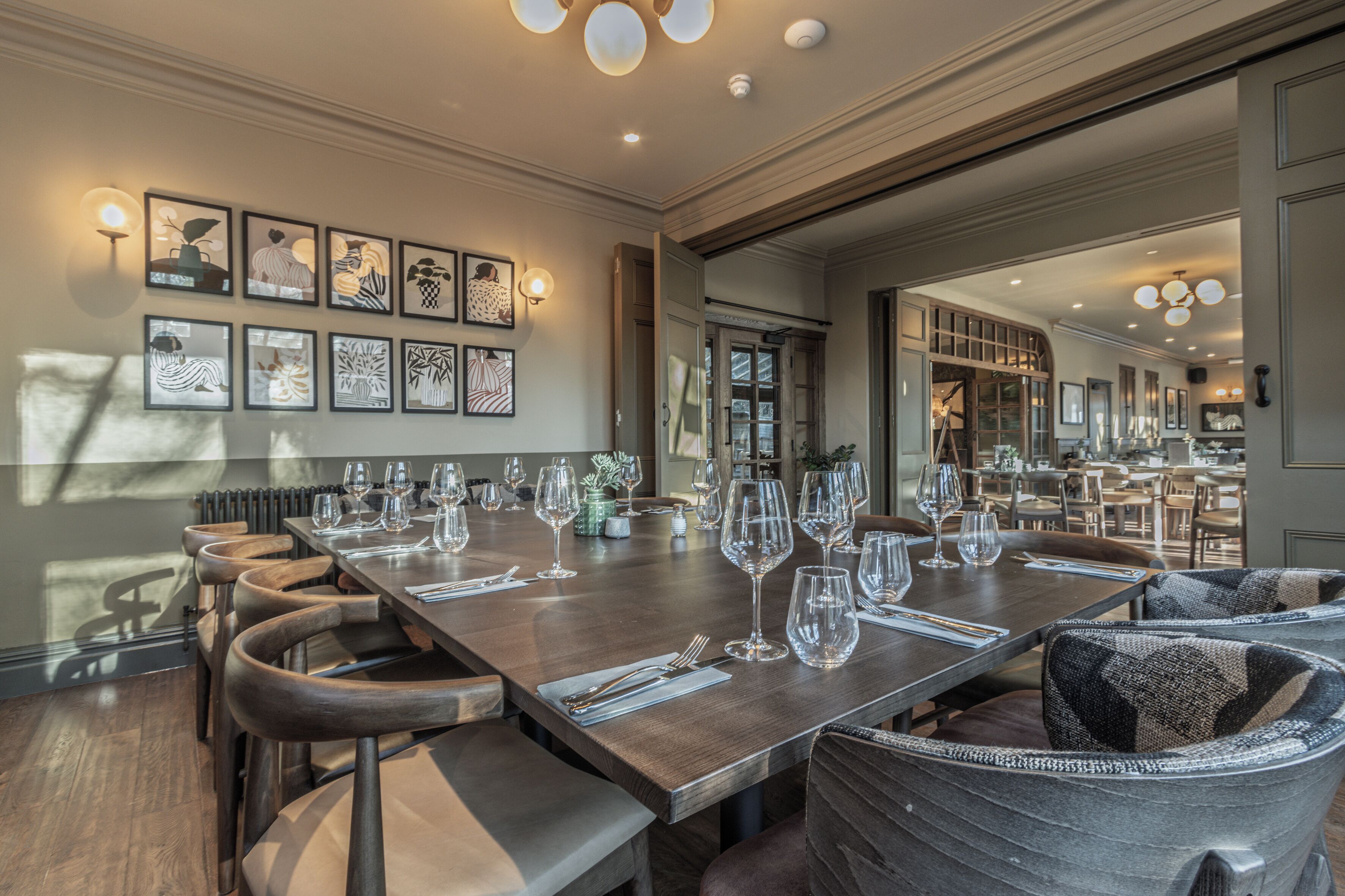 A dining area with a long table set for multiple guests, framed artwork on the wall, and a view into the kitchen.