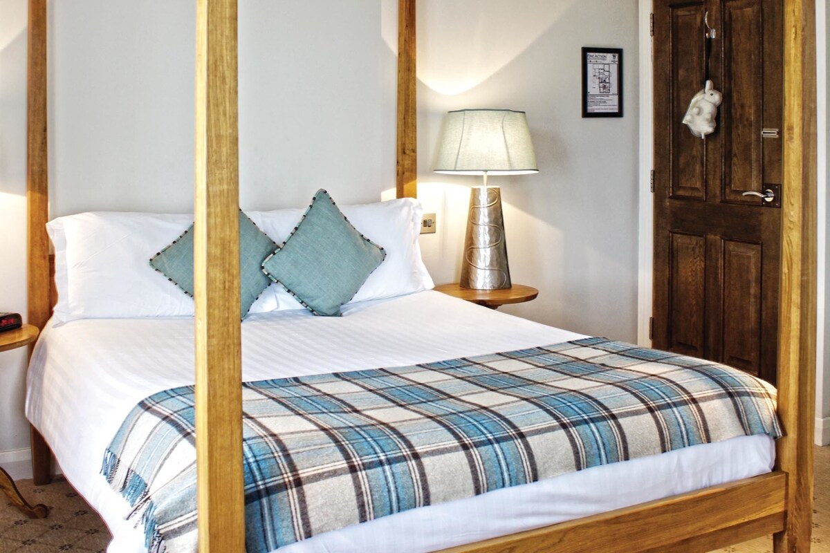 A four-poster bed with a plaid bedspread, a bedside lamp, and a wooden door in a room with patterned carpet.