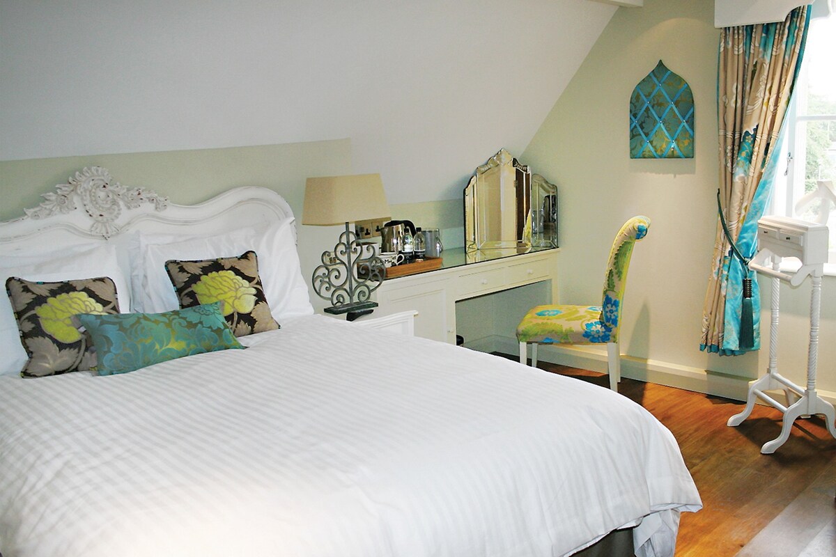 A bedroom with a large bed, a desk, a chair, a mirror, and a window with curtains.