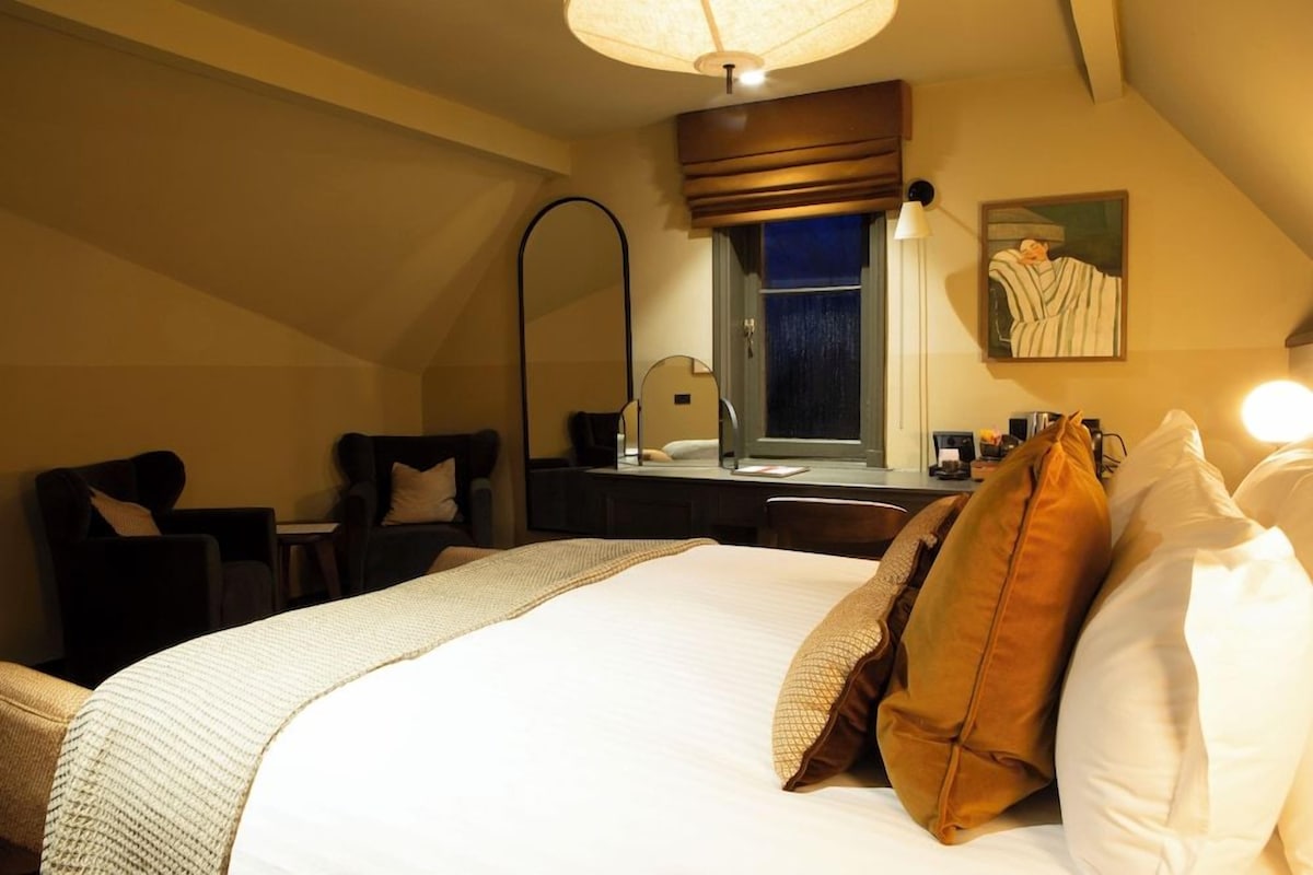 A hotel room with a large bed, a desk, a chair, a lamp, and a painting on the wall.