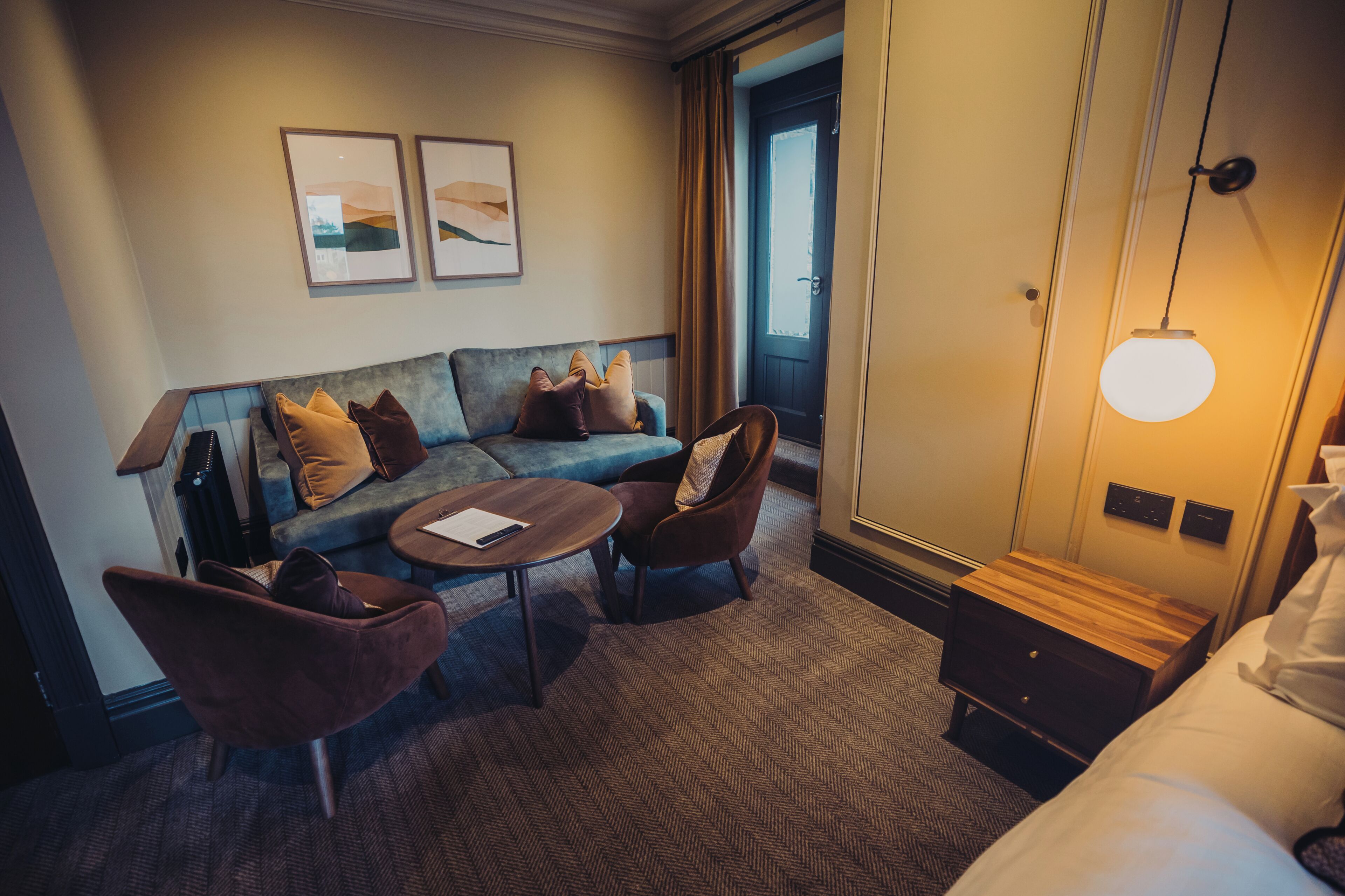 A hotel room with a sofa, armchairs, a small table, a bed, a nightstand, and a lamp.