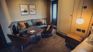 A hotel room with a sofa, armchairs, a small table, a bed, a nightstand, and a lamp.