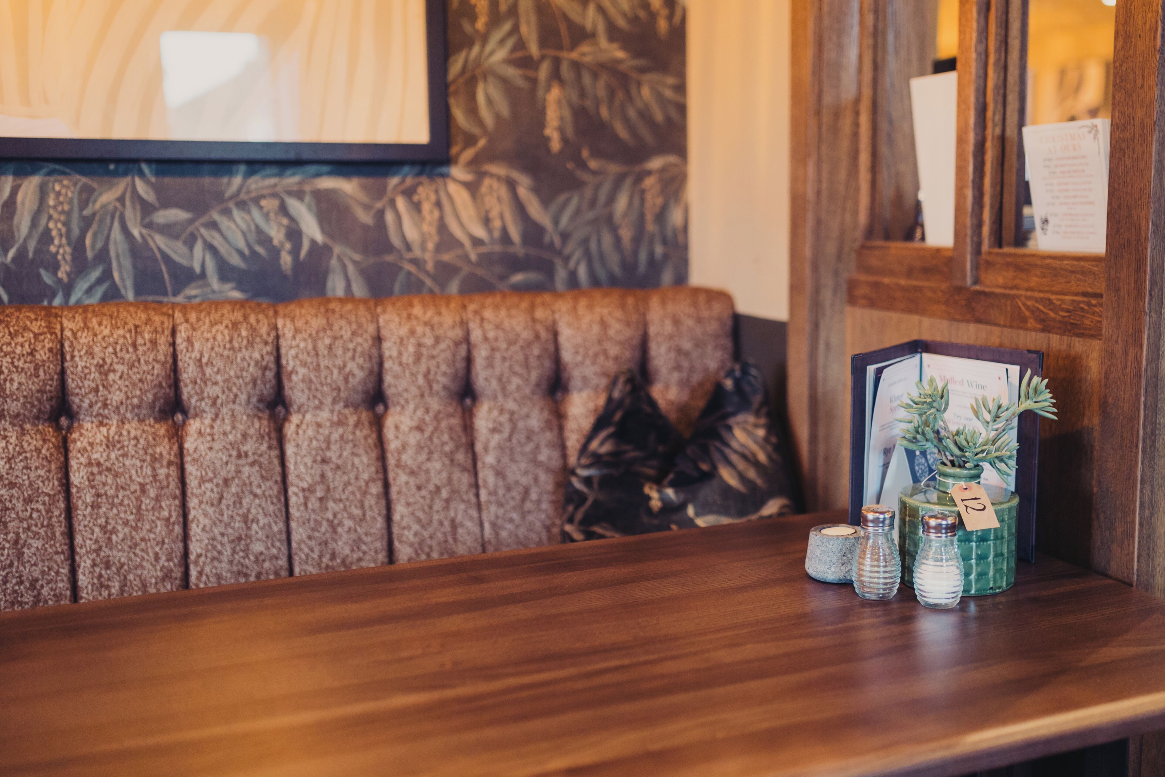 A wooden bench with a tufted backrest, a framed picture of a succulent plant, and three glass bottles with labels on a wooden table.