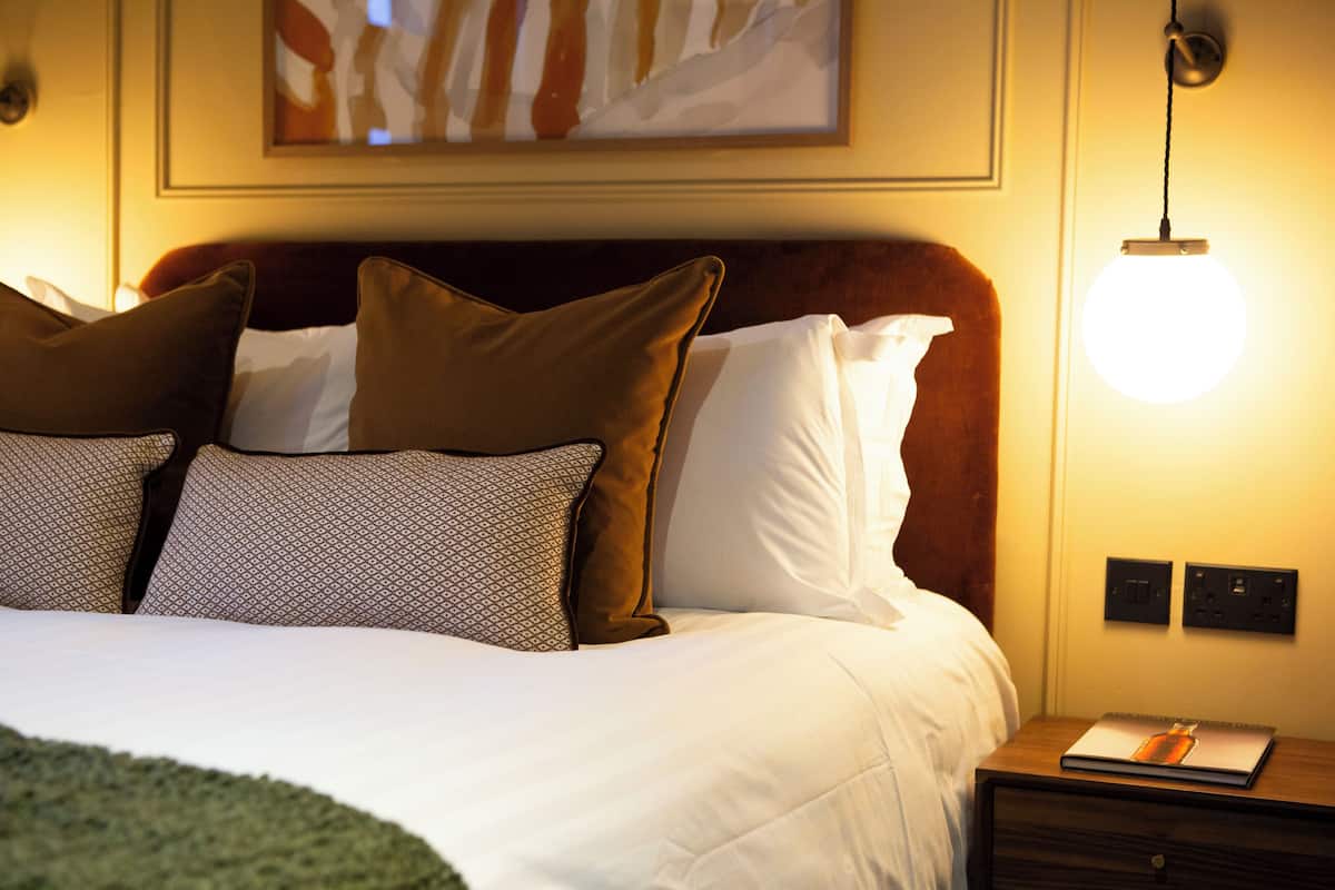 A neatly made bed with patterned and solid pillows, a wooden headboard, a bedside lamp, and a small table with a notebook.