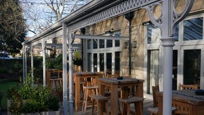 An outdoor patio with wooden tables and chairs, a pergola, and a stone wall. - The Wheatley Arms (Ilkley)