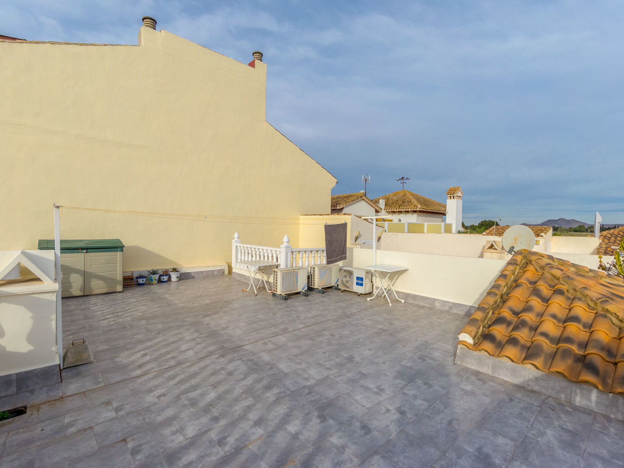 Una terraza en la azotea con barandilla blanca, una mesa y una silla. Se observan plantas en macetas y una antena parabólica.
