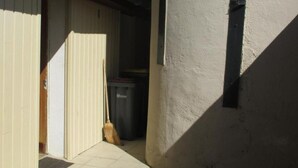 Un étroit couloir extérieur au sol carrelé, avec un balai appuyé contre le mur et une poubelle en arrière-plan.