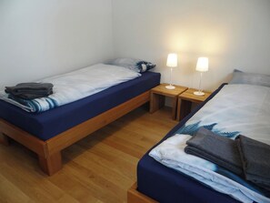 A small, compact room with two single beds, each with a wooden frame and blue bedding. There are two bedside tables with lamps, and the room has a wooden floor.