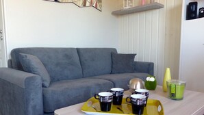 Un salon avec un canapé gris, une petite table sur laquelle se trouve un plateau avec des tasses à café, une figurine de grenouille verte et un vase jaune.