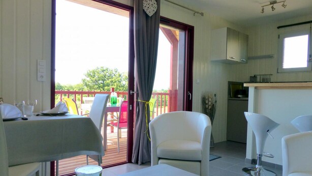 Une pièce comprenant une table à manger, deux chaises blanches, un petit coin cuisine et une porte-fenêtre coulissante donnant sur une terrasse extérieure.