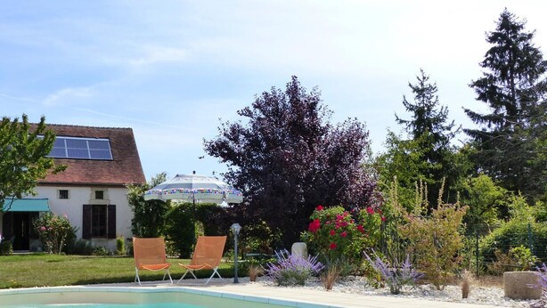 Une piscine à l’eau d’un bleu limpide, entourée d’un jardin où poussent diverses plantes et fleurs, et une maison avec des panneaux solaires en arrière-plan.