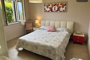 A bedroom with a bed, a nightstand, a lamp, a painting, and a window. - Villa Bella Vista—Panoramic Sea View near Tropea (Zambrone)
