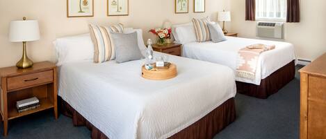A hotel room with two beds, a nightstand with a lamp, a small shelf with books, and framed pictures on the wall.