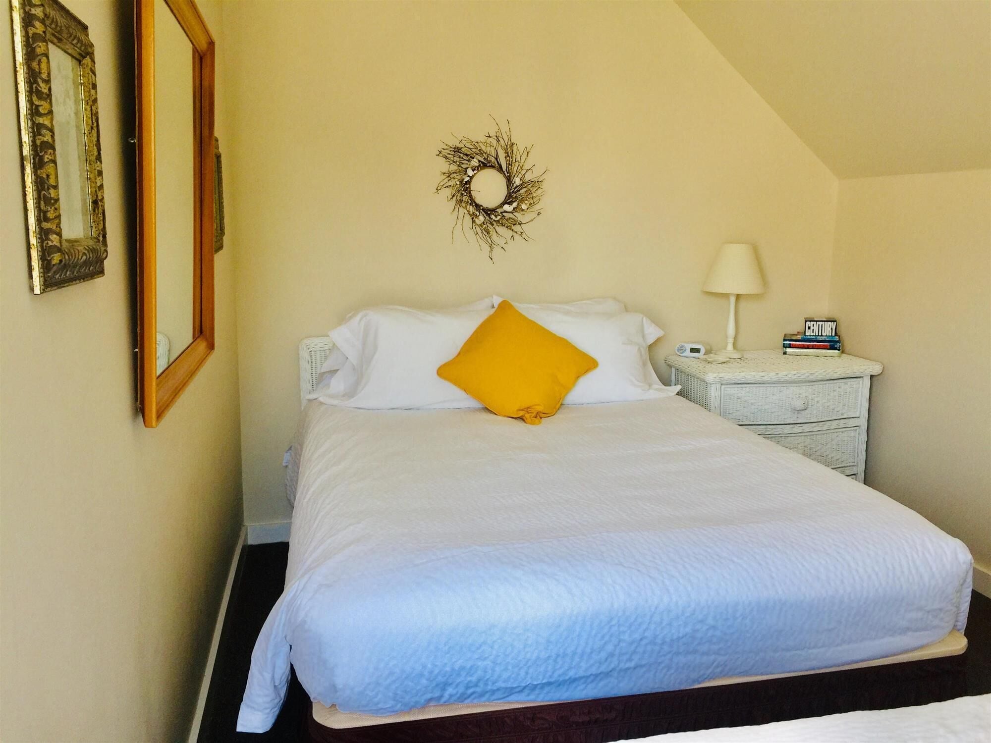 Un lit bien fait, avec une literie blanche et bleue, un oreiller jaune, une couronne décorative accrochée au mur, une table de chevet avec une lampe et une petite horloge.